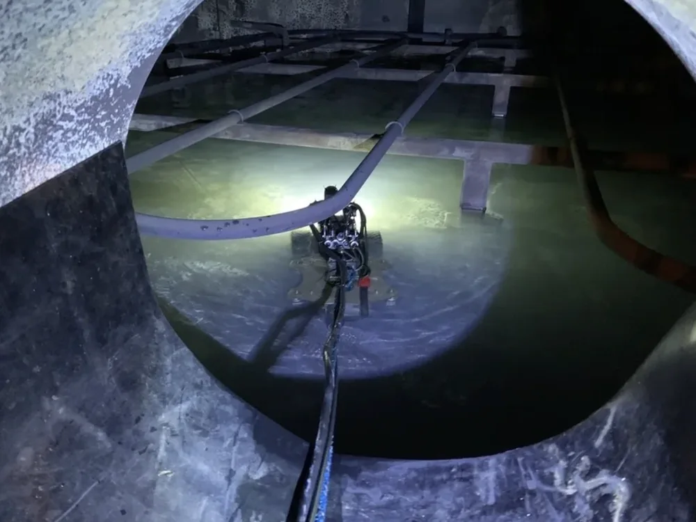 A submersible crawler lit by its own lighting on the floor of a water tank, seen through the manway while the tank remains full.